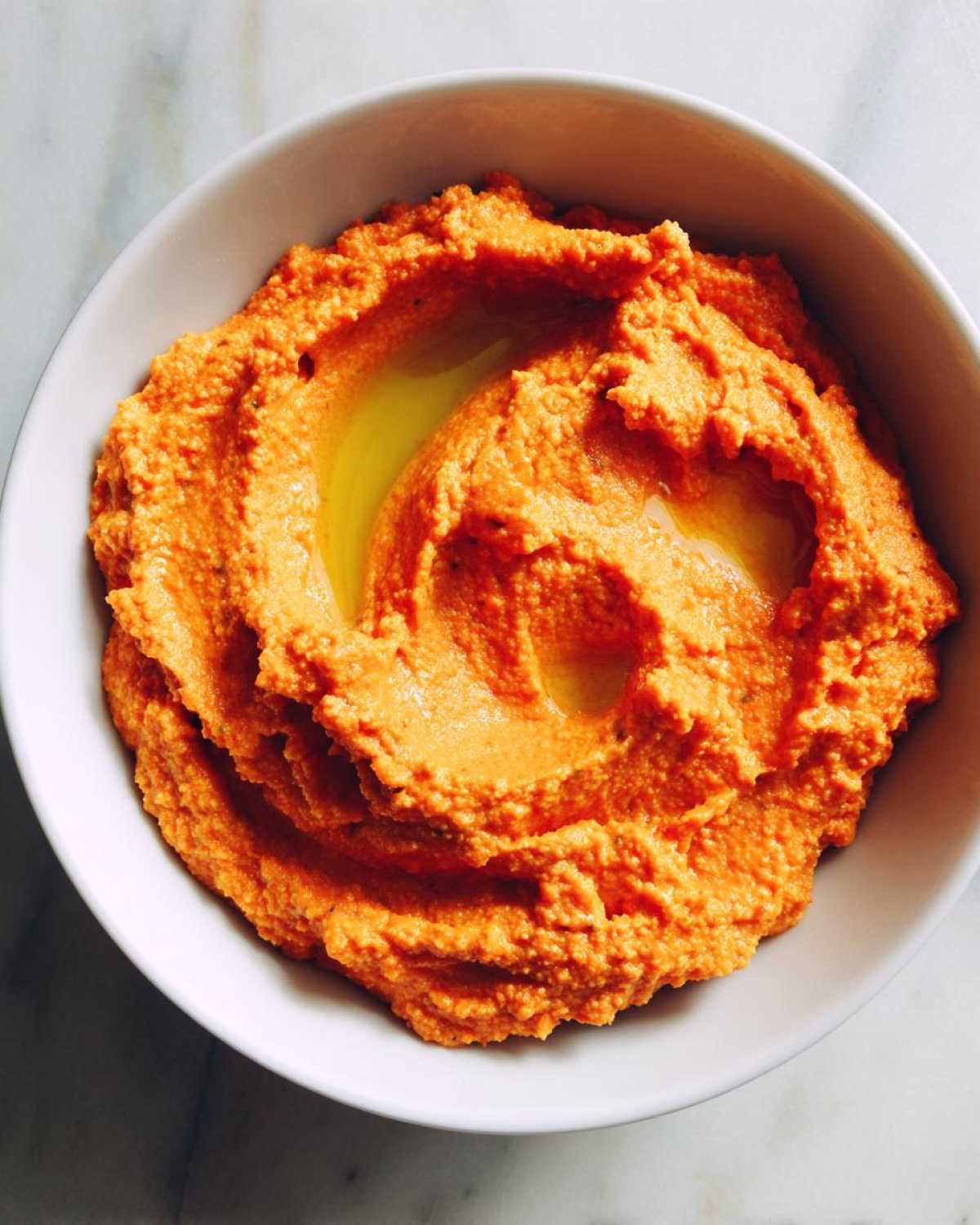 Overhead view of vibrant orange Roasted Red Pepper Hummus swirled in a white bowl, topped with olive oil.