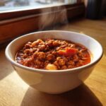 A steaming white bowl filled with rich, red Spicy Turkey White Bean Chili, sitting on a wooden surface.