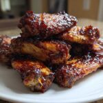 A stack of crispy oven baked Sticky BBQ Chicken Wings glistening with dark, rich sauce on a white plate.