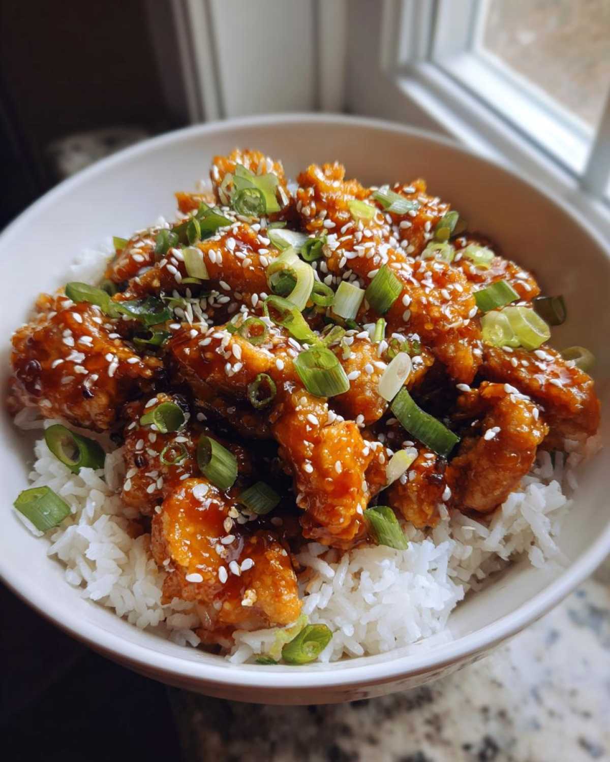 Close-up of a Sticky Orange Chicken Bowl served over white rice, topped with sesame seeds and chopped green onions.