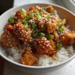 Close-up of a Sticky Orange Chicken Bowl with rice, topped with sesame seeds and chopped green onions.
