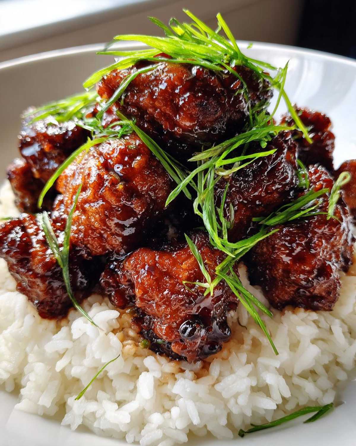 Close-up of Sticky Teriyaki Chicken Bites coated in a dark, glossy sauce, served over a bed of fluffy white rice and garnished with green onions.