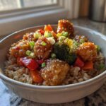 A close-up of a Sweet Chili Chicken Bowl featuring glazed chicken pieces over brown rice with broccoli, peppers, and sesame seeds.