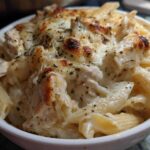 A close-up of a serving of Cheesy Chicken Alfredo Bake with Mozzarella, featuring penne pasta, chicken pieces, and a bubbly, browned cheese topping.