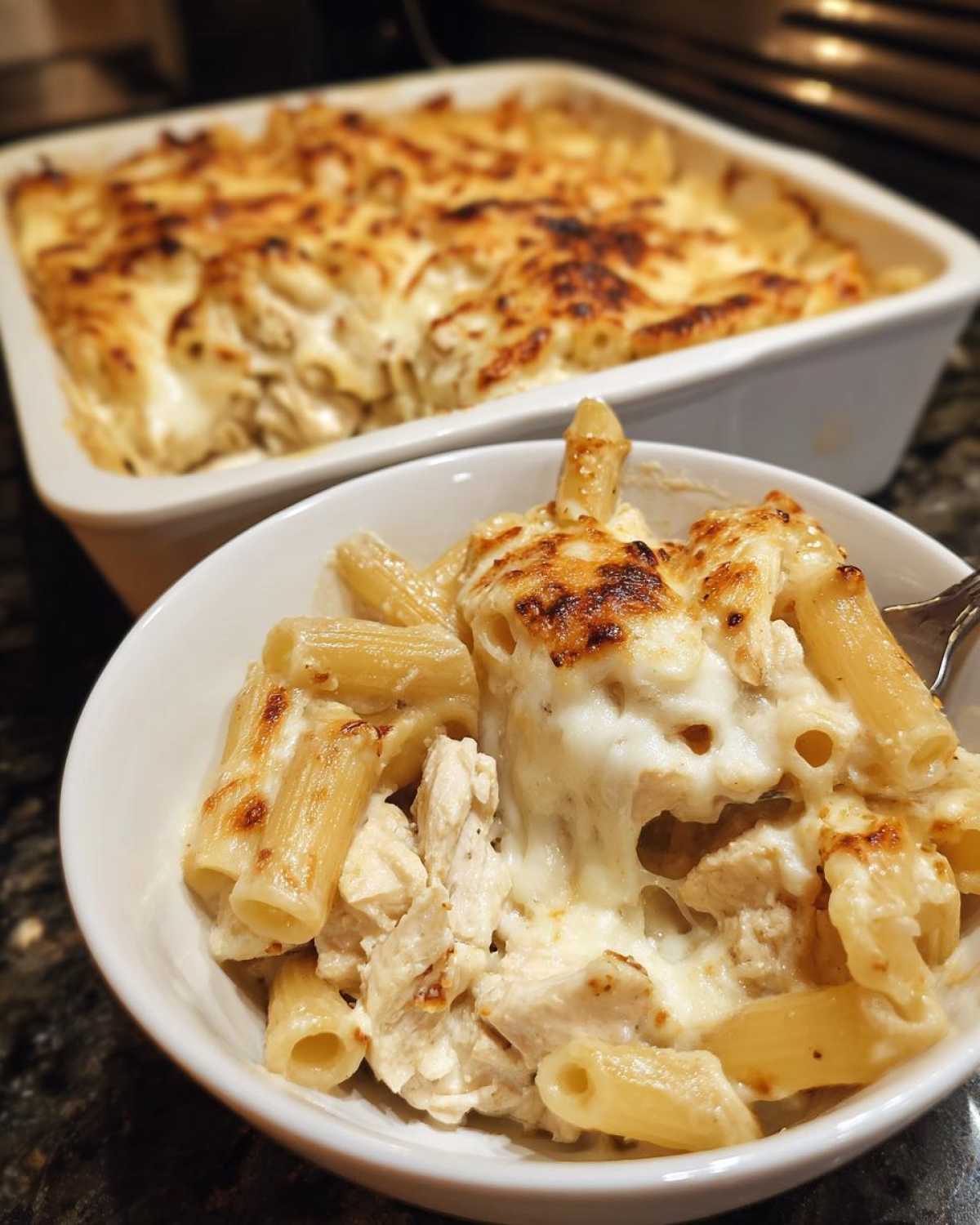 A close-up bowl of Cheesy Chicken Alfredo Bake with Mozzarella showing melted, browned cheese pull over pasta and chicken.