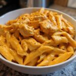 A close-up of a white bowl filled with Cheesy Chicken Queso Pasta, featuring penne pasta coated in a vibrant orange, creamy sauce with chunks of seasoned chicken.