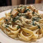 A close-up of rich Creamy Spinach Alfredo Pasta with Chicken served over fettuccine noodles on a white plate.