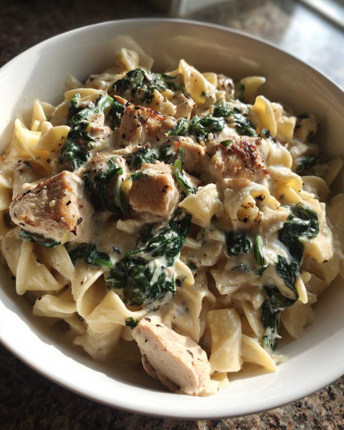 A close-up of Creamy Spinach Chicken Pasta with Garlic served in a white bowl, featuring chunks of chicken and wilted spinach.
