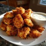A pile of golden brown, crispy chicken nuggets served with a small ramekin of creamy garlic dip.