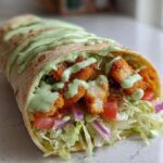 Close-up of a Crispy Chicken Wrap filled with lettuce, tomato, onion, and topped with a generous drizzle of green avocado sauce.