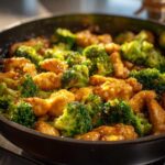 Close-up of Garlic Butter Chicken and Broccoli Stir Fry coated in a glossy sauce cooking in a dark skillet.