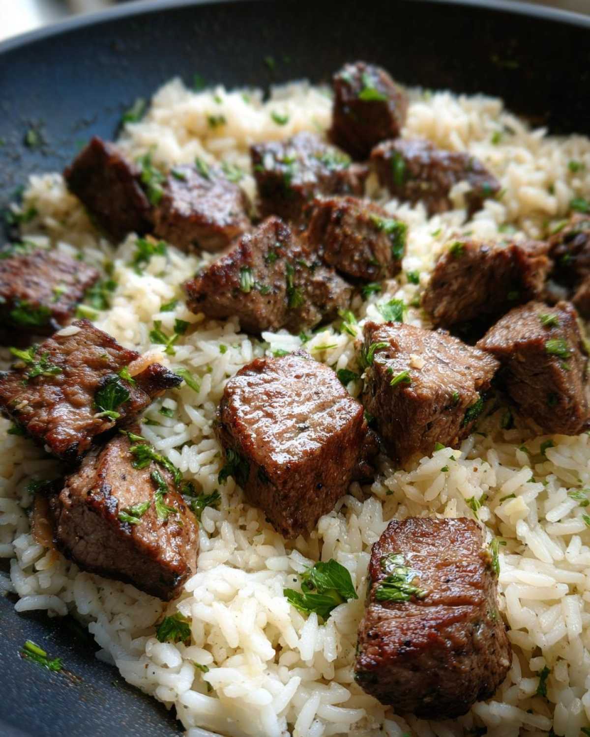 Close-up of juicy steak cubes served over white rice, garnished with parsley in a skillet for Garlic Butter Steak and Rice Skillet Dinner.