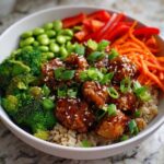 A close-up of a Honey Garlic Chicken Bowl featuring glazed chicken pieces over brown rice, surrounded by broccoli, edamame, carrots, and scallions.