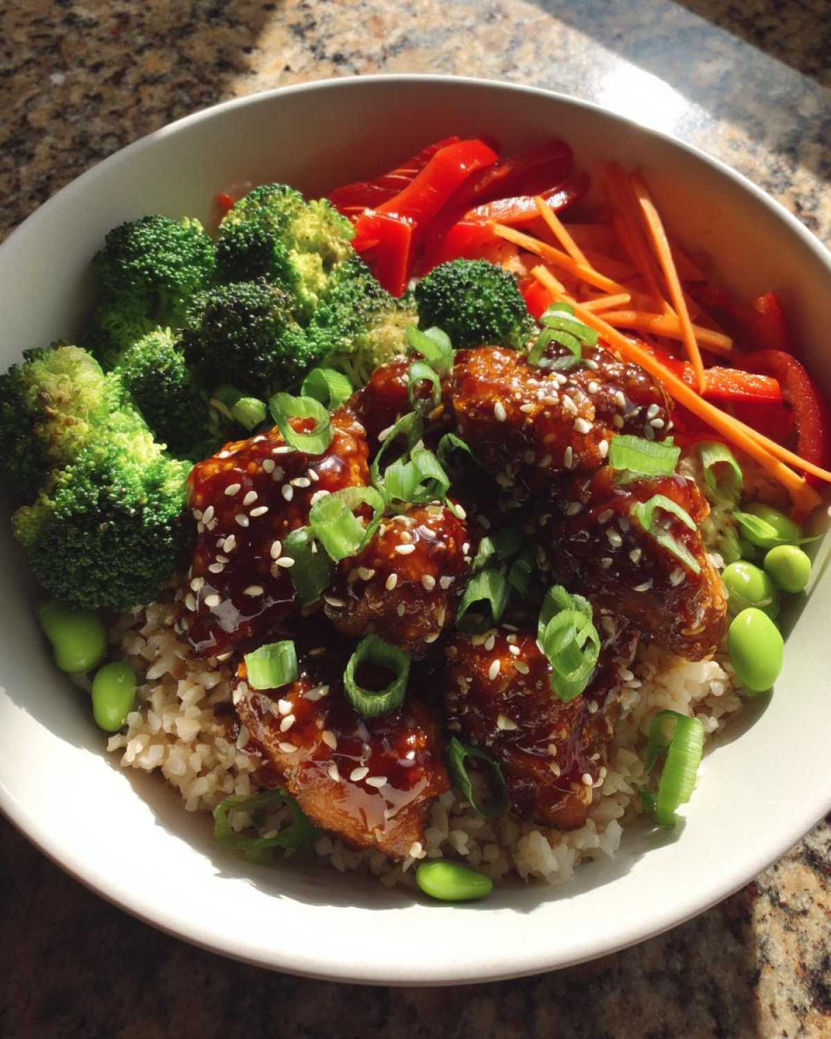 Close-up of a Honey Garlic Chicken Bowl featuring glazed chicken pieces over brown rice, surrounded by broccoli, peppers, carrots, and edamame.
