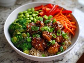A close-up of a Honey Garlic Chicken Bowl featuring glazed chicken pieces over brown rice, surrounded by broccoli, edamame, carrots, and scallions.