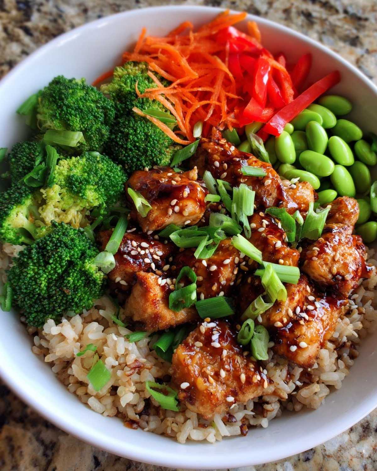 Close-up of a Honey Garlic Chicken Bowl featuring glazed chicken pieces over brown rice, topped with broccoli, edamame, carrots, and green onions.