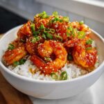 A white bowl filled with white rice topped with glossy Spicy Honey Garlic Shrimp, garnished with sesame seeds and sliced green onions.
