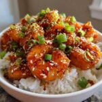 Close-up of a white bowl filled with white rice topped with glossy Spicy Honey Garlic Shrimp, garnished with sesame seeds and green onions.