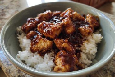 Close-up of a bowl filled with white rice topped generously with glazed, sticky BBQ chicken pieces.