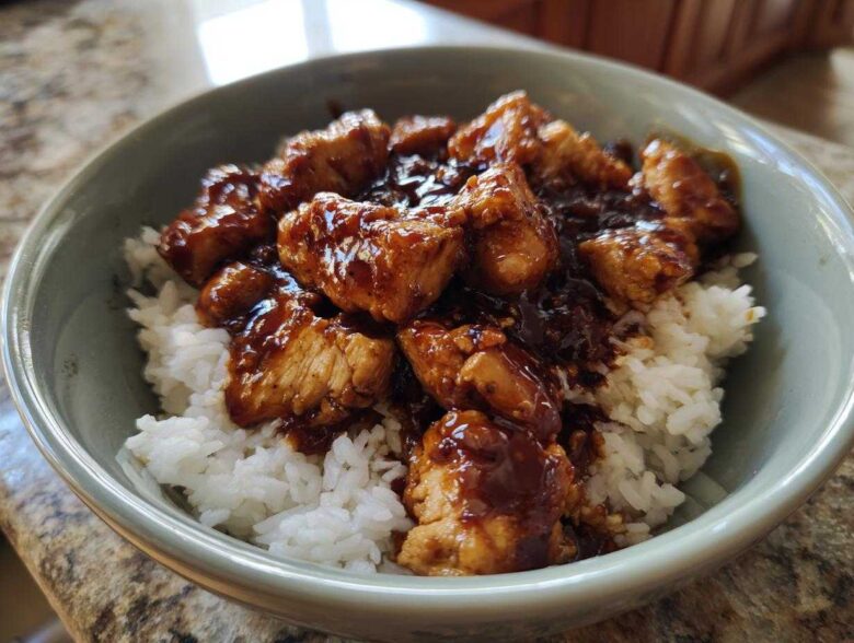 Close-up of a bowl filled with white rice topped generously with glazed, sticky BBQ chicken pieces.